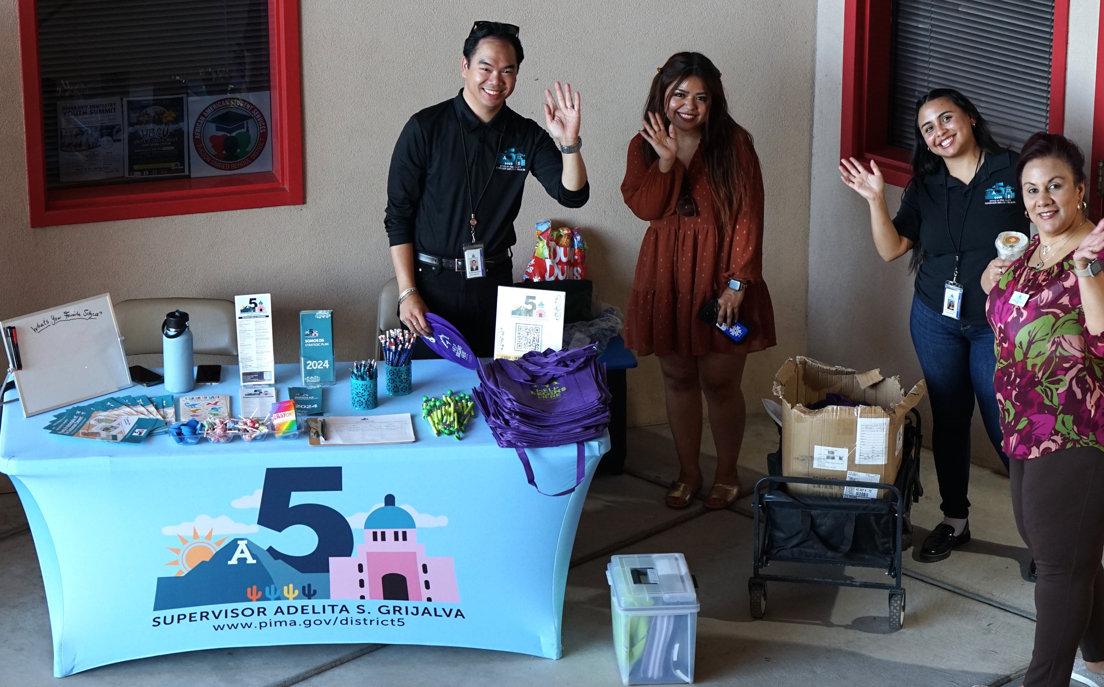 Four employees smile and wave from Supervisor Adelita Grijalva's table at the fair