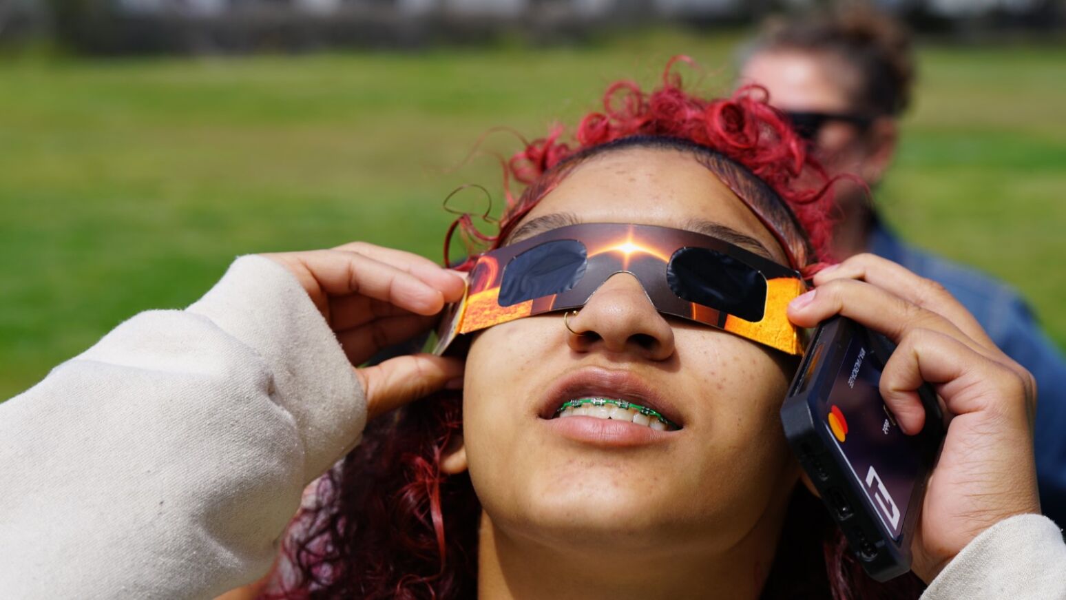 A teen girl with curly red hair looks at the eclipse through her special glasses