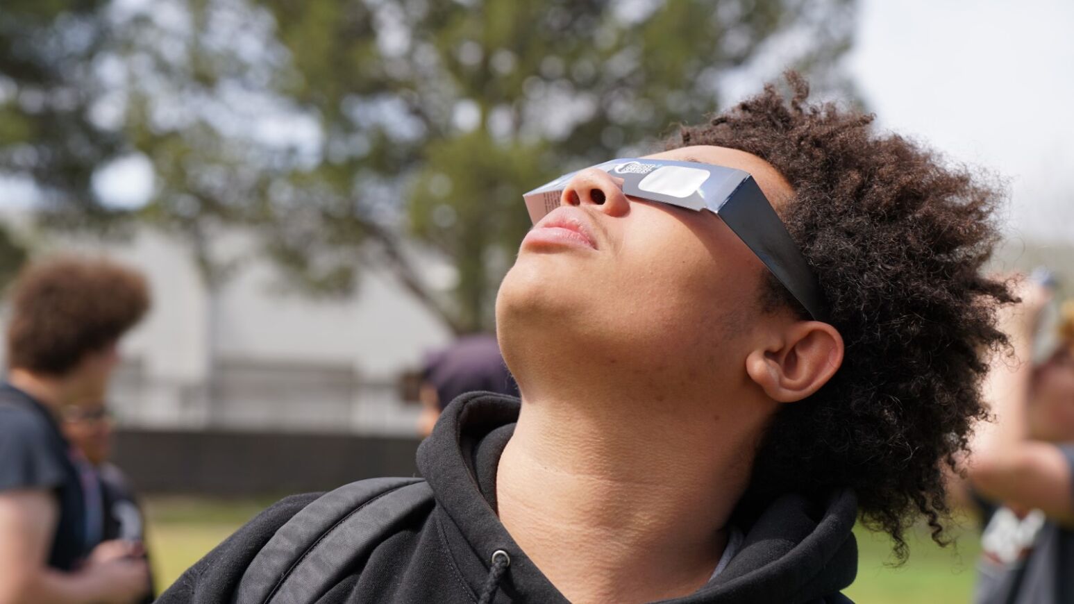 A teen boy with curly hair looks up at the eclipse with his special glasses